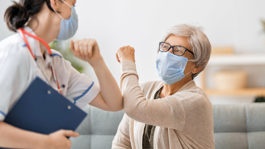 Cheerful older woman bumping elbows with healthcare worker. Both are wearing masks.