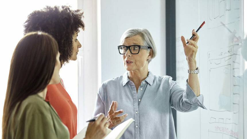 Senior female entrepreneur explaining strategy on glass wall to businesswomen. Colleagues are discussing in board room. They are working in office.