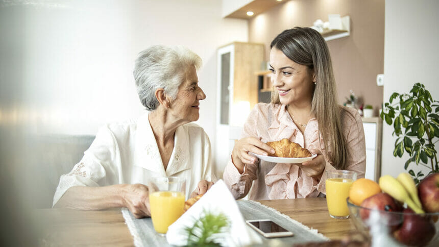 Cheerful young girl serving breakfast to an elderly woman at home - A social worker visiting an elderly woman