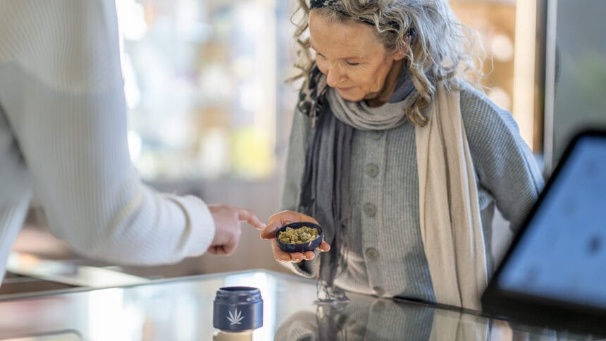 A mature woman stands at the counter of a legal cannabis retailer as she holds out a lid of buds. She is dressed casually and looking down to inspect the product as the associate tries to sell it to her.