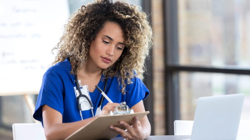 Young mixed race female nurse writes something on a clipboard while preparing for a weekly staff meeting. An open laptop is on the table.