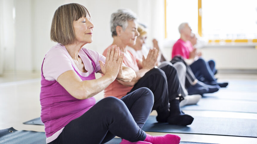 Relaxed senior woman meditating with friends in yoga class. Elderly females and males are exercising while sitting in row at gym. They are in sportswear.