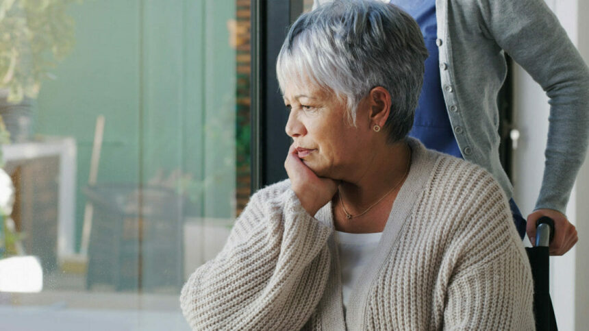 older women in wheelchair pensively looking out window. Aide behind her.