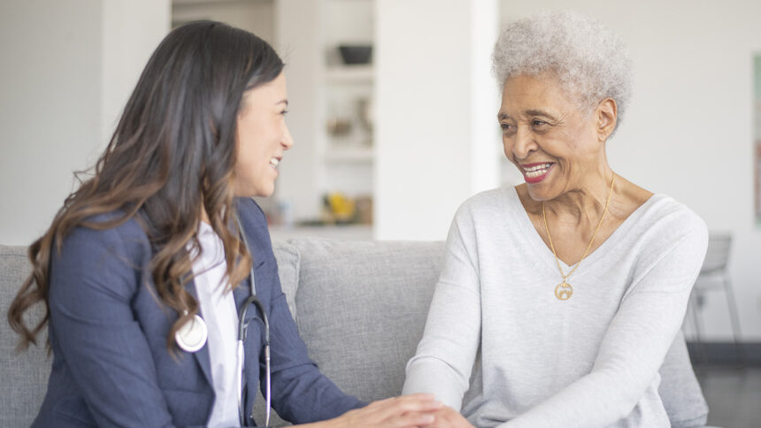 A female doctor makes a house call to a senior female patient. They are sitting together on the sofa as they discuss her health concerns and how she can support her patient.