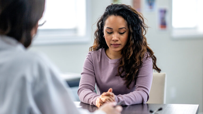 A young woman of mixed race, sits with her female doctor as they discuss her mental health. The patient is dressed casually and appears depressed as she looks down low with a forlorn expression on her face.