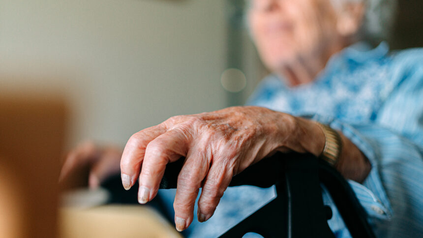 Close-Up Focus on Foreground Shot of an Unrecognizable Senior Woman Resting Her Hand on the Grip of her Mobility Walker while Sitting and Looking Out the Window