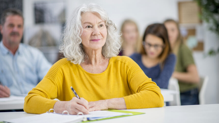 A diverse group of students are indoors in a university. They are taking night classes. A Caucasian senior woman is in front, and she is smiling while watching the lecture.