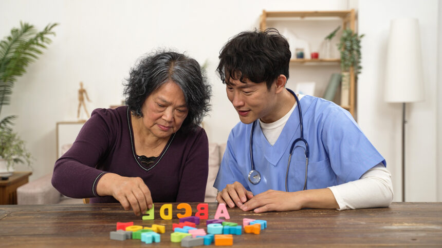 asian male physiotherapist applauding for elderly Parkinson’s patient as she is trying to arrange color letter blocks with hand tremor. in-home care and rehabilitation for senior concept