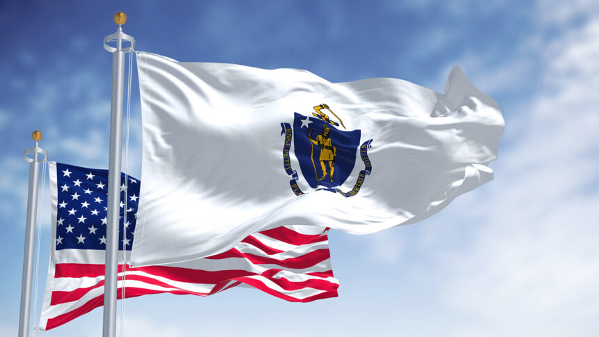 The Massachusetts state flag waving along with the national flag of the United States of America. In the background there is a clear sky. Massachusetts is a state in the New England region of the United States